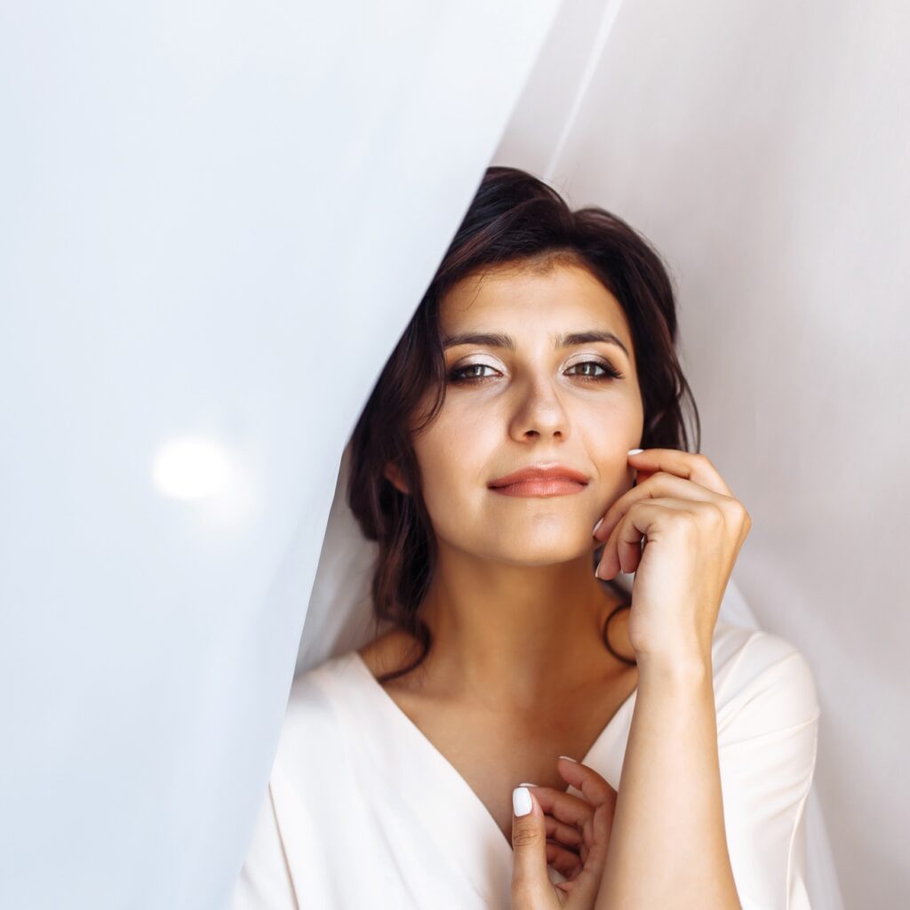 Elegant bride in white robe posing under curtain. Amazing bride in the morning preparation.